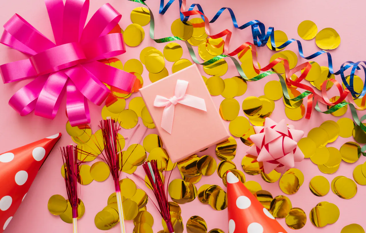 DIY-Dekoideen für eine unvergessliche Feier: Kreative Tipps und Tricks Rosa Geschenkbox mit Schleife, goldenem Konfetti, Partyhüten, Partybläsern und bunten Luftschlangen auf einem rosa Hintergrund - perfekte Dekoideen für jede Feier.