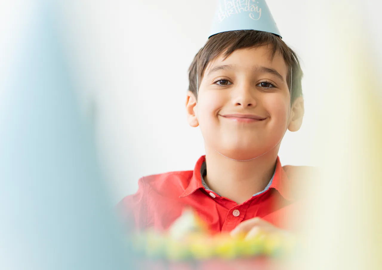 10 kreative Geburtstagsthemen für Kinder: Tolle Ideen für unvergessliche Feiern Ein lächelndes Kind mit rotem Hemd und hellblauem "Happy Birthday"-Hut sitzt an einem Tisch, umgeben von verschwommenen Objekten und festlichen Geburtstagsthemen.