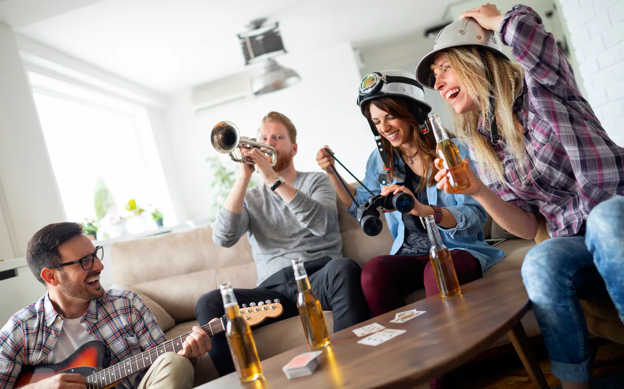 5 lustige Geburtstagsspiele für jedes Alter: Spaß für alle Gäste Vier Erwachsene versammeln sich in einem Wohnzimmer und spielen Geburtstagsspiele; einer spielt Gitarre, ein anderer Trompete, zwei tragen Helme und halten Ferngläser. Auf dem Tisch stehen Spielkarten und Flaschen, während sie gemeinsam Getränke genießen.