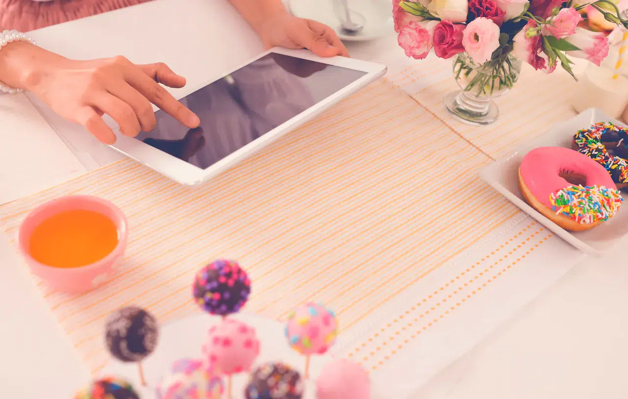 DIY-Dekoideen für eine unvergessliche Feier: Kreative Tipps und Tricks Eine Person benutzt ein Tablet auf einem mit Dekoideen gedeckten Tisch, darunter eine Tasse mit orangefarbener Flüssigkeit, bunte Donuts, Cake Pops und eine Vase mit rosa Blumen.