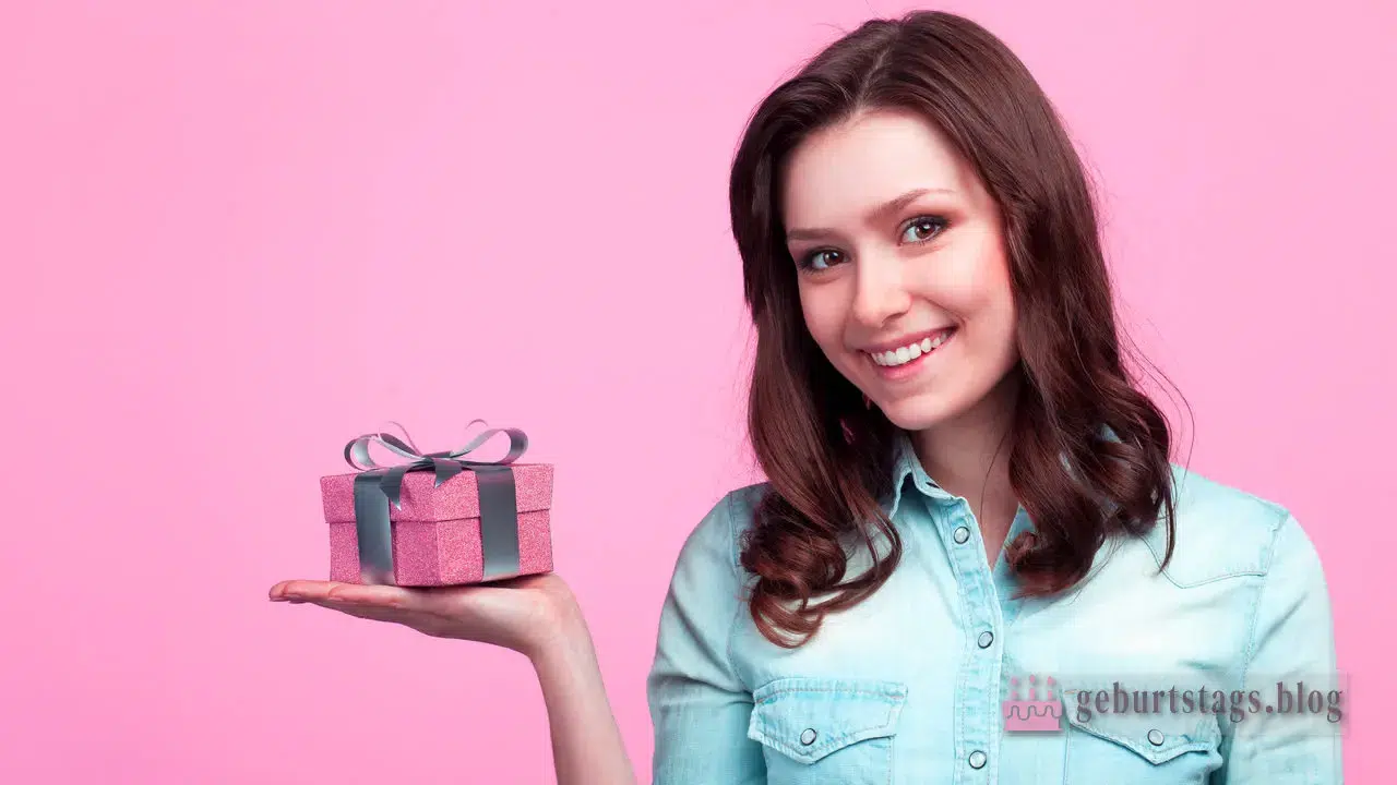 Lächelnde Frau im Jeanshemd hält eine kleine rosa Geschenkbox mit Schleife vor rosa Hintergrund, perfekt für kreative Überraschungen und Geschenkideen für Frauen.