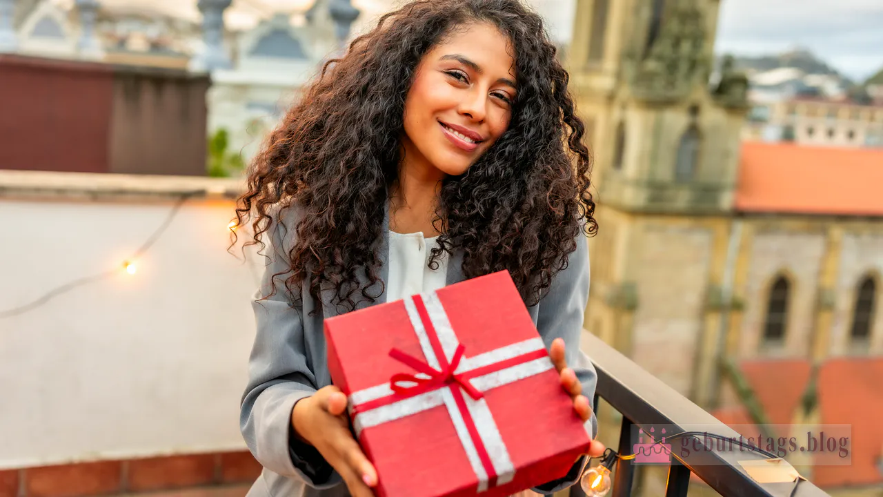 Eine Person mit lockigem Haar auf einem Balkon hält lächelnd eine rote Geschenkbox mit einer weißen Schleife – eine der perfekten Geschenkideen, um Ihre Freundin zu überraschen.