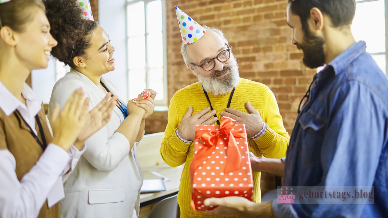 Eine Gruppe von Menschen mit bunten Partyhüten feiert einen Geburtstag in einem Büro. Eine Person hält ein großes rotes Geschenk mit Punkten in der Hand, was zu den Überraschungen des Tages beiträgt.