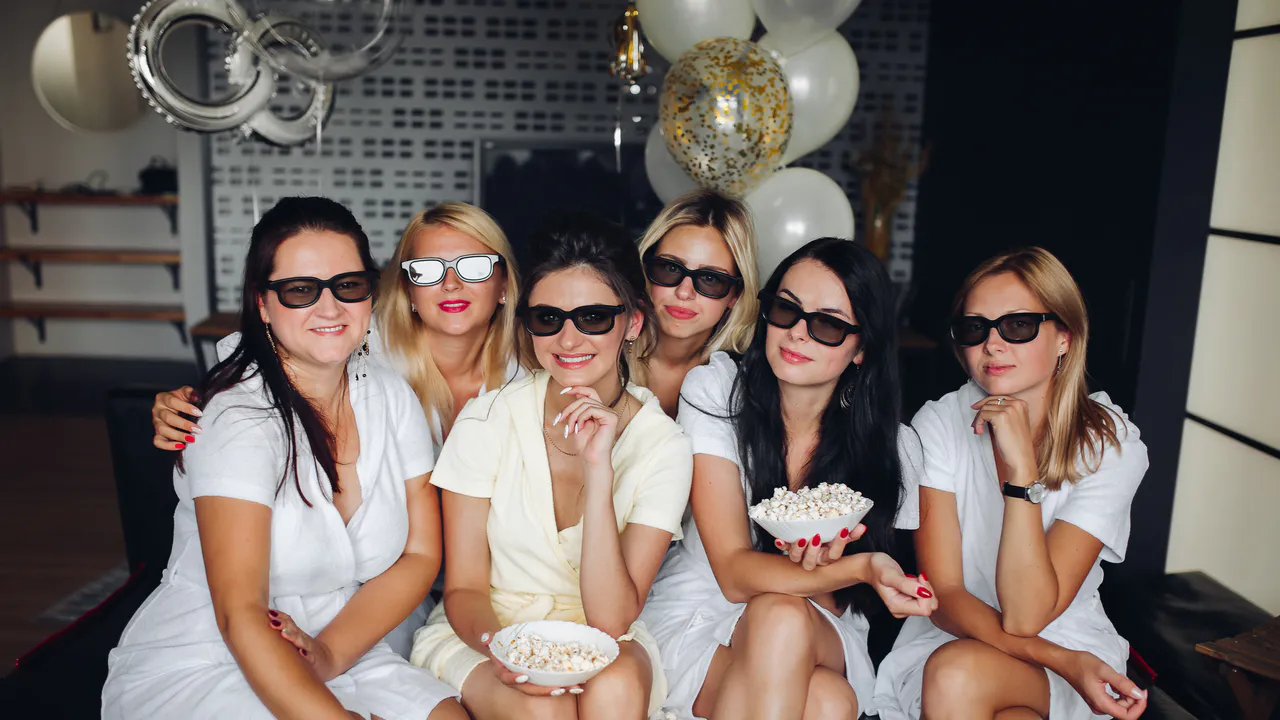 Sechs Frauen mit 3D-Brillen und hellen Outfits sitzen zusammen drinnen, halten Popcorn-Schalen, Ballons im Hintergrund - eine perfekte Inspiration dafür, wie du eine Glamour-Geburtstagsparty gestaltest.