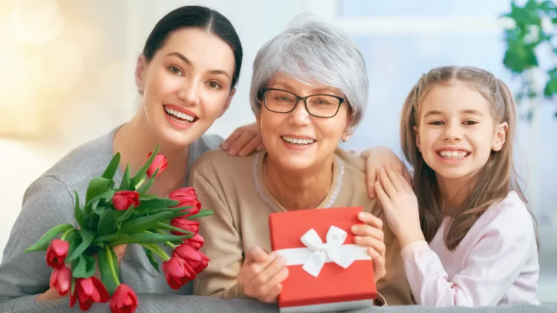 Drei Generationen von Frauen lächeln; die Älteste hält ein Geschenk mit weißem Band, die Jüngste umarmt sie, und die Dritte trägt rote Tulpen - perfekte Geschenkideen für Mama: Liebevolle Präsente für die beste Mutter.
