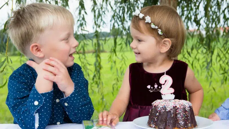 Ein Junge und ein Mädchen sitzen an einem Tisch im Freien mit einem Schokoladen-Geburtstagskuchen, der mit einer Kerze mit der Nummer 2 verziert ist - eine Szene, die Geschenkideen für den 2. Geburtstag: niedlich und praktisch inspiriert.