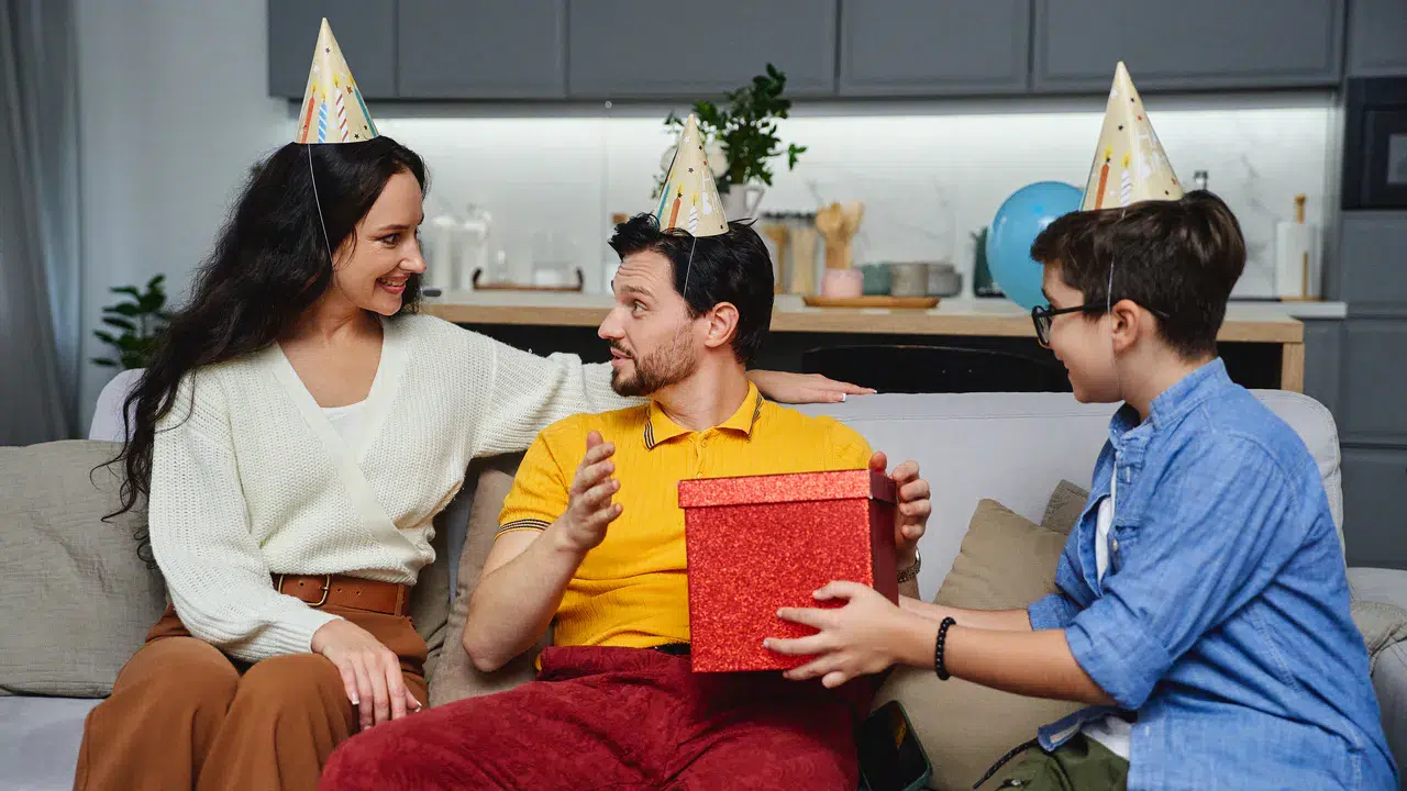Drei Personen mit Partyhüten sitzen auf einer Couch in einer häuslichen Umgebung; eine überreicht eine rote Geschenkbox - eine Inspiration für Geburtstags Geschenkideen: Passende Präsente für jedes Alter - und alle lächeln und genießen den Moment.