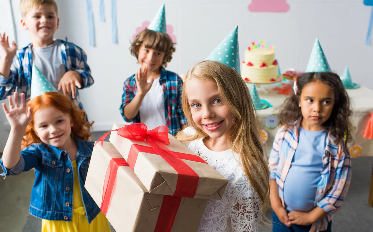 Fünf Kinder auf einer Geburtstagsparty tragen Partyhüte; ein Mädchen im Vordergrund hält zwei eingepackte Geschenke mit roten Bändern, die tolle Geburtstags Geschenkideen präsentieren: Passende Präsente für jedes Alter. Kuchen und Dekoration vervollständigen die festliche Szene.