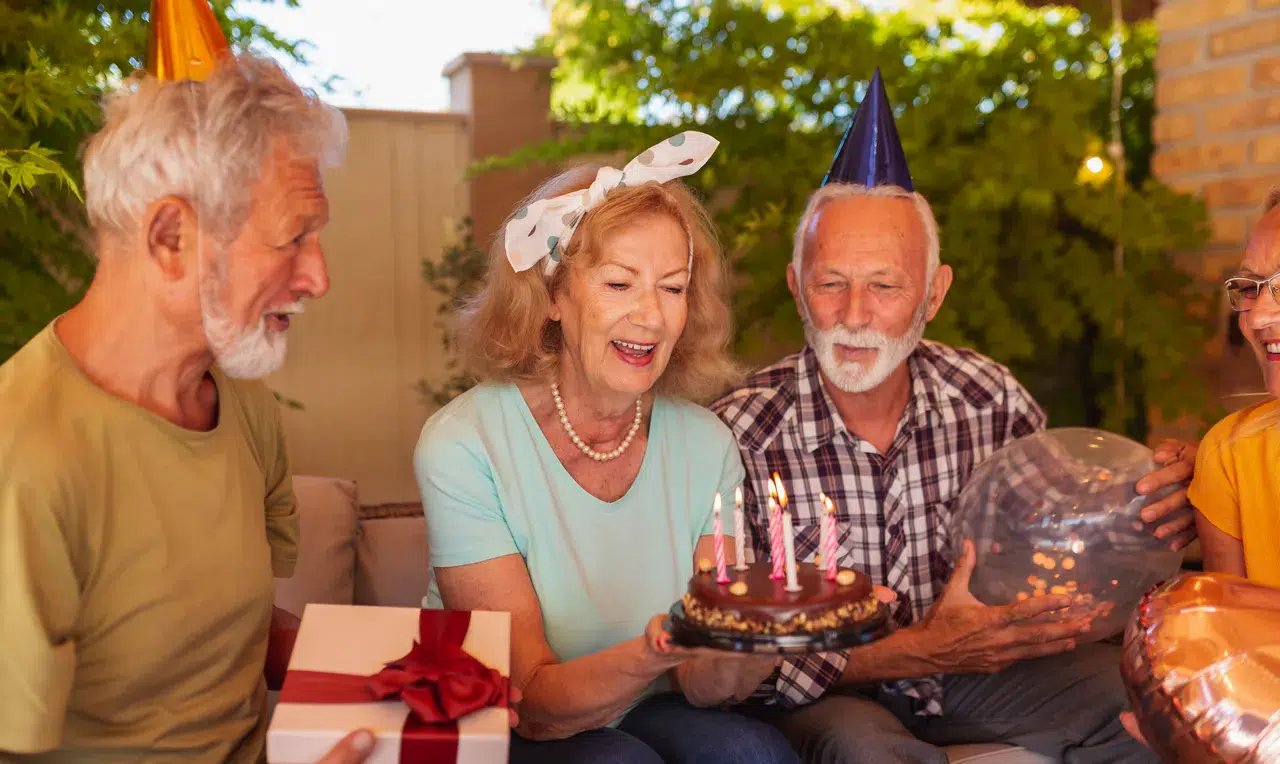 Vier ältere Erwachsene auf einer Party im Freien, die Partyhüte tragen. Eine Frau hält eine Geburtstagstorte mit brennenden Kerzen, während ein anderer Mann Geburtstags-Geschenkideen überreicht: Passende Präsente für jedes Alter schön verpackt für die Feier.