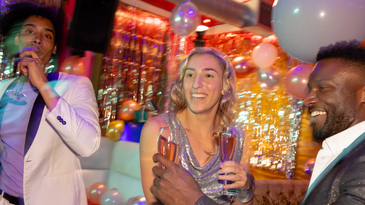 Drei Personen in Partykleidung halten Sektgläser und lächeln, umgeben von Luftballons und bunten Dekorationen - die perfekte Inspiration für Wie du eine Glamour-Geburtstagsparty gestaltest.