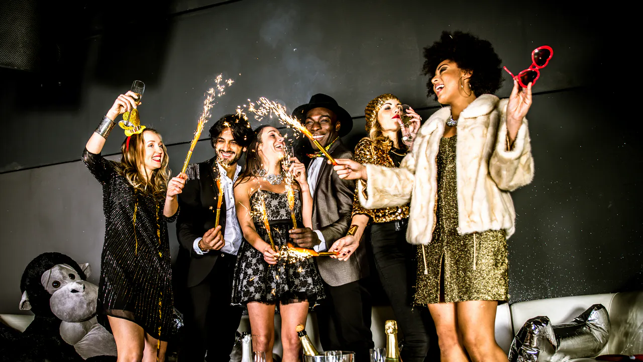 Sechs Personen in festlicher Kleidung feiern zusammen drinnen, halten Wunderkerzen und Getränke-Partydeko im Hintergrund zeigt, wie du eine Glamour-Geburtstagsparty gestaltest.