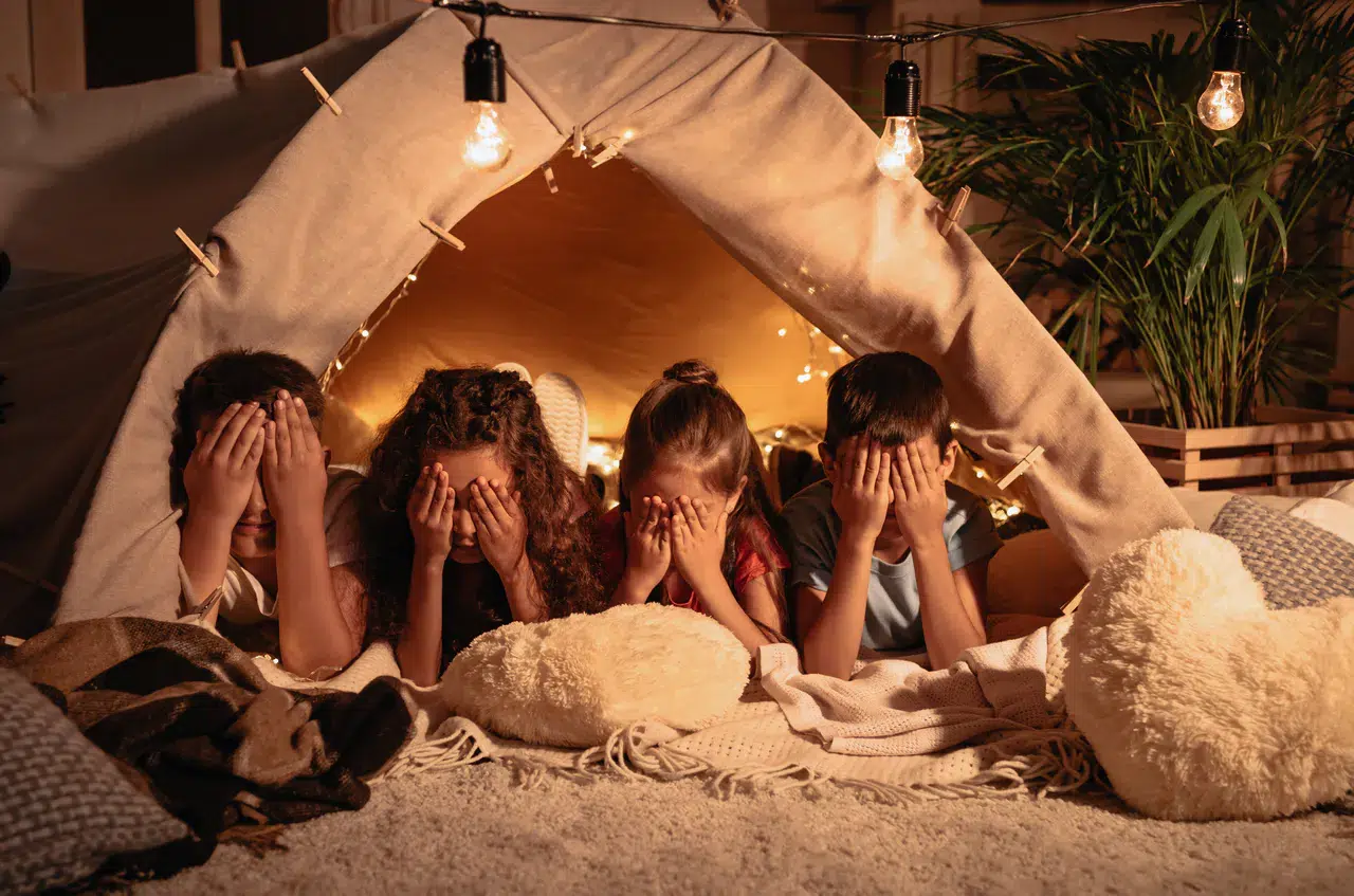 Vier Kinder, die unter einer Zeltdecke mit Lichterketten liegen und ihre Gesichter mit den Händen bedecken, umgeben von Kissen und gemütlicher Dekoration - perfekt für Geburtstagsfeiern im Herbst: Ideen und Inspirationen für unvergessliche Momente.