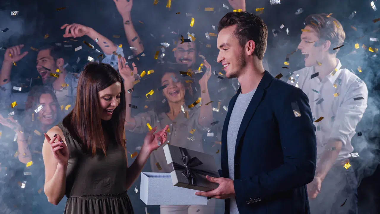 Ein Mann überreicht einer lächelnden Frau auf einer Party eine Geschenkbox, während Konfetti fällt und die Leute feiern - so gelingt ein unvergesslicher Moment, wenn du eine Glamour-Geburtstagsparty gestaltest.