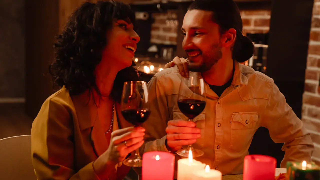 Geschenkideen für die Freundin: Romantisch und kreativ Eine Frau und ein Mann sitzen eng beieinander, lächelnd und mit Rotweingläsern in der Hand, auf dem Tisch vor ihnen stehen brennende Kerzen - eine Szene voller Geschenkideen für Freundin: Romantisch und kreativ.