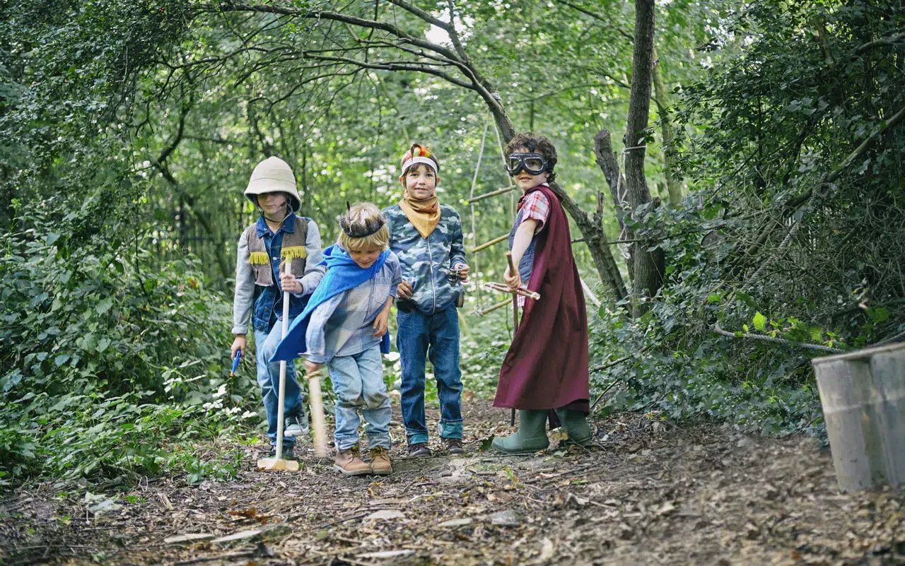 Vier Kinder in Kostümen spielen mit Holzstöcken auf einem Waldweg - umgeben von Bäumen und Laub. Spaßige Geburtstagsspiele für draußen machen ihre gemeinsame Zeit unvergesslich.