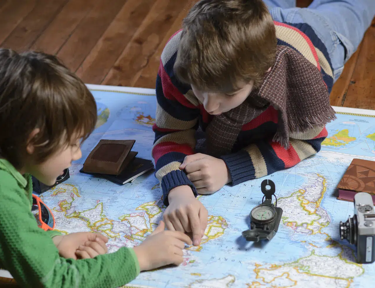Zwei Kinder liegen auf dem Boden und schauen sich gemeinsam eine Karte an - mit Kompass, Kamera und Notizbüchern beim Planen einer abenteuerlichen Schatzsuche.