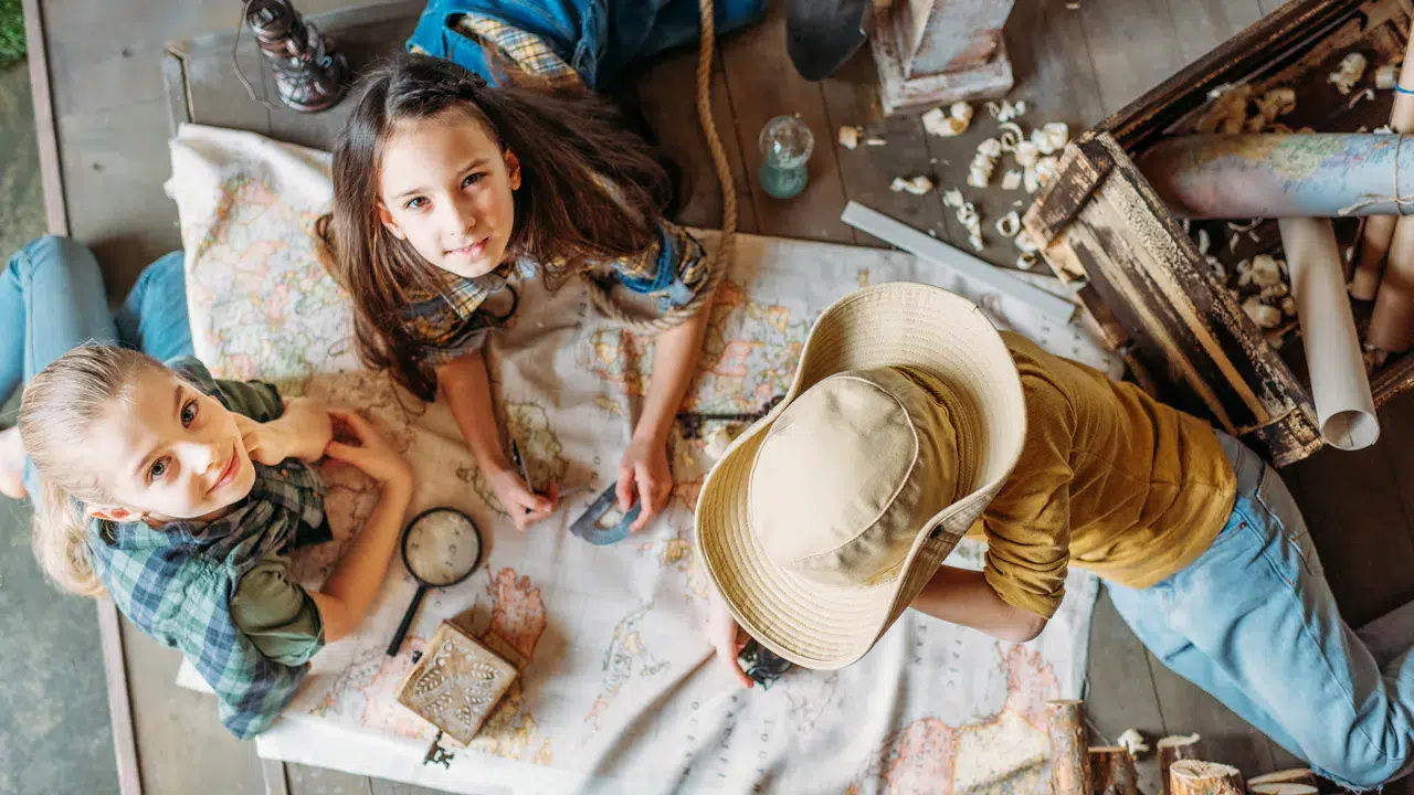 Drei Kinder sitzen auf dem Boden, umgeben von einem Kompass, einer Lupe und einer offenen Holzkiste voller kleiner Schätze - sie sind vertieft ins Planen einer abenteuerlichen Schatzsuche mit einer großen Karte vor sich.