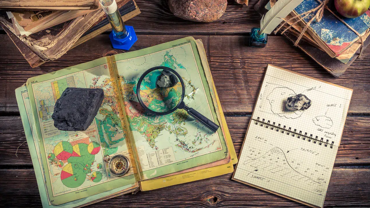Eine offene Weltkarte mit Lupe, Felsen, Kompass, Notizbuch mit Skizzen und alten Büchern auf einem Holztisch, perfekt für Planen einer abenteuerlichen Schatzsuche oder eine immersive Geologie- oder Geografiestudienszene.