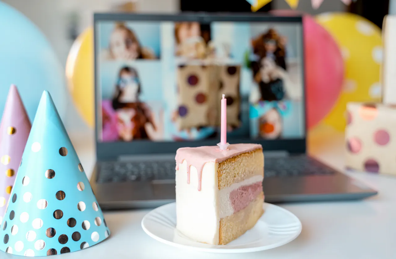 Ein Stück Schichtkuchen mit einer brennenden Kerze liegt auf einem Teller vor einem Laptop, auf dem ein Videoanruf und Partyhüte angezeigt werden, und fängt den Geist von Geburtstagsfeiern im Herbst ein: Ideen und Inspirationen zum Feiern online.