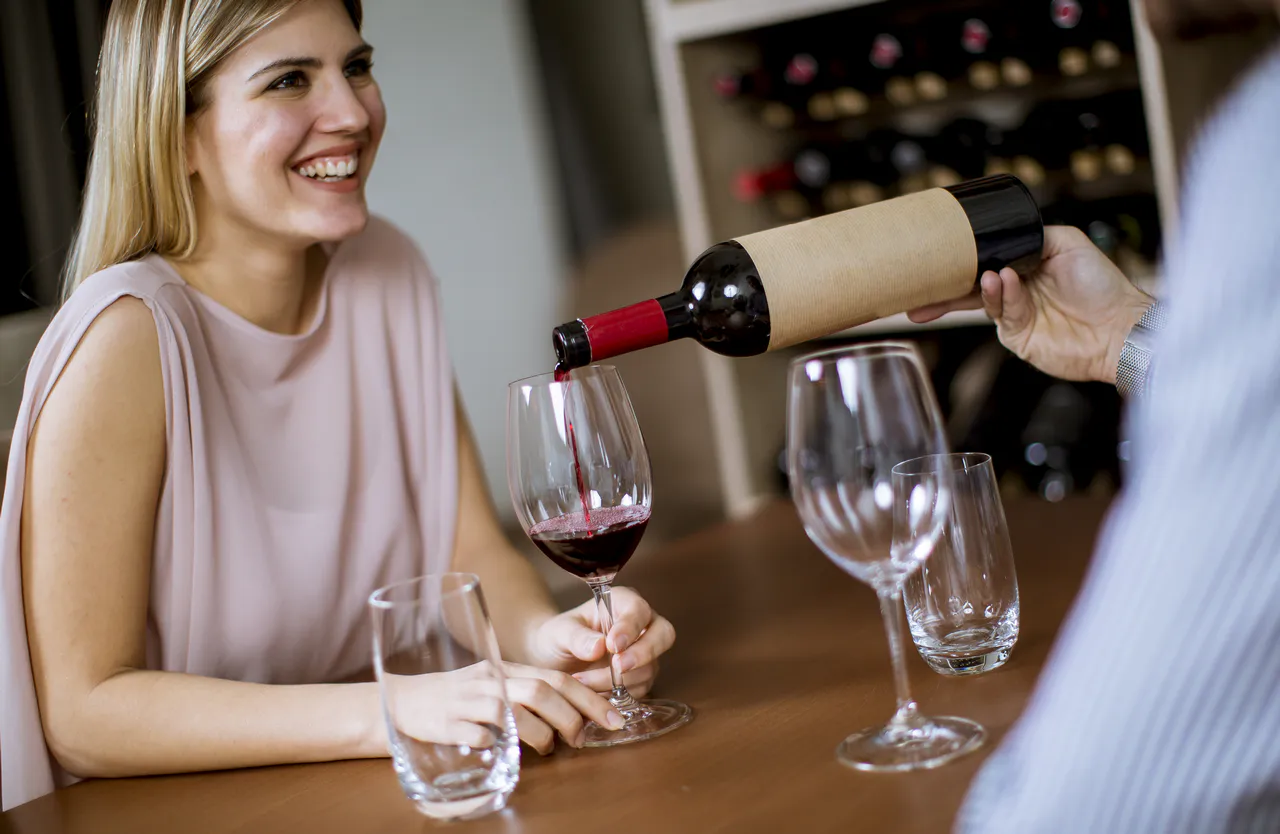 Eine Person gießt Rotwein in ein Glas für eine lächelnde Frau, die an einem Tisch sitzt, umgeben von leeren Gläsern und Weinflaschen - perfekt für Geburtstagsfeiern im Herbst: Ideen und Inspirationen.